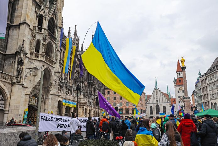 Zahlreiche Menschen haben sich eingefunden zur Ukraine-Kundgebung „Gemeinsam gegen den Krieg – ein Jahr des großen Krieges“ am 24. Februar 2023 auf dem Marienplatz. Eine große Ukraineflagge weht über der Menschenmenge.
