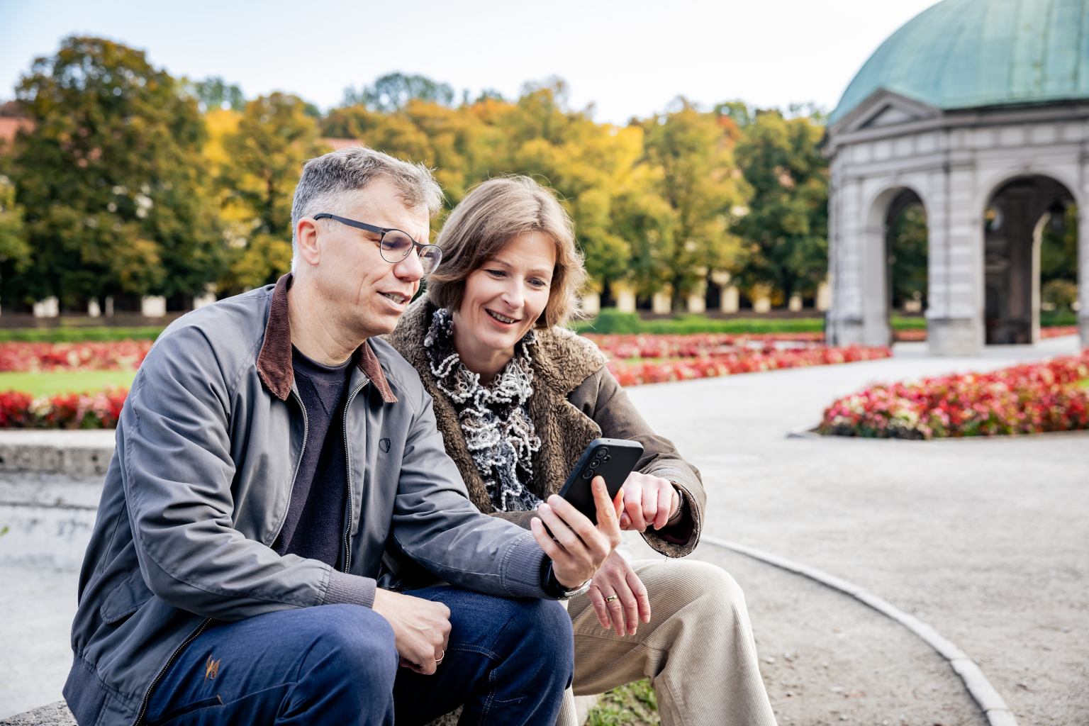 Mann und Frau sitzen auf Brunnenrand im Münchner Hofgarten, schauen auf Handy, Pavillon im Hintergrund