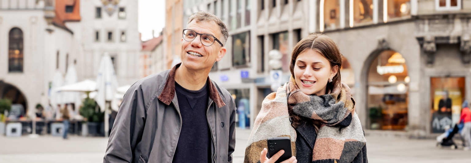 Mann und Frau auf dem Marienplatz. Mann schaut zum Rathausturm, Frau schaut aufs Handy