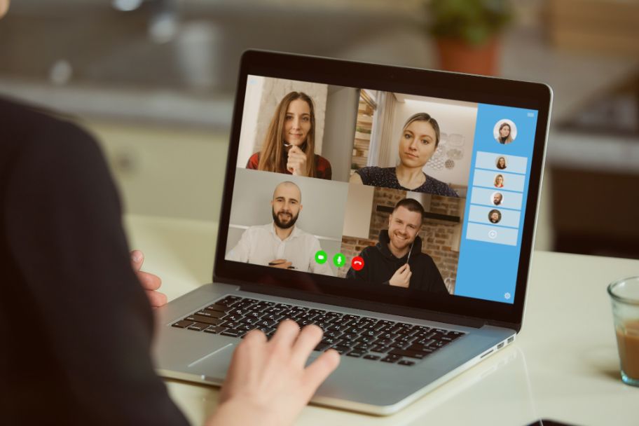 Foto eines Notebook-Bildschirms mit 4 Gesichtern, Kamera blickt einem weiteren Teilnehmer oder Teilnehmerin über die Schulte