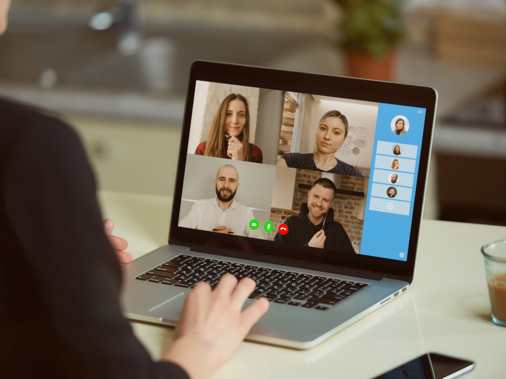 Foto eines Notebook-Bildschirms mit 4 Gesichtern, Kamera blickt einem weiteren Teilnehmer oder Teilnehmerin über die Schulte